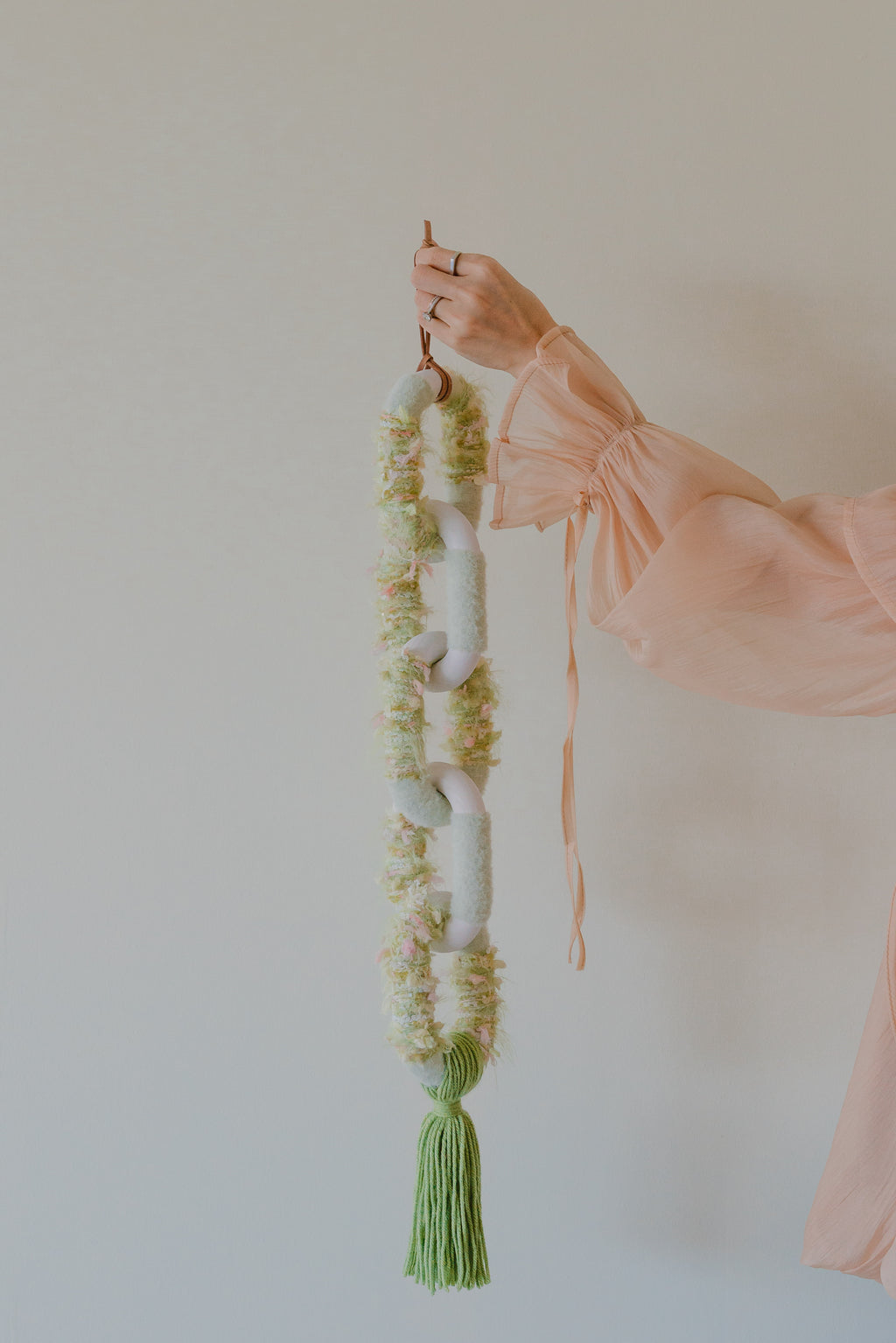 Passing By Blooms Valley fibre wall art — layered yarn in soft sage and cream tones, woven with leather accents for gentle floral texture.
